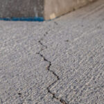 A visible crack runs through the surface of a concrete pavement near the corner of a building.
