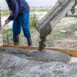 Worker leveling freshly poured concrete from a truck chute onto a driveway or foundation area outdoors.