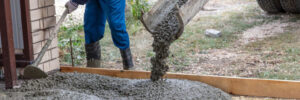 Worker leveling freshly poured concrete from a truck chute onto a driveway or foundation area outdoors.