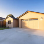 Single-story beige house with a two-car garage, concrete driveway, and fenced backyard under a clear sky.