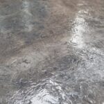 Wet, textured concrete patio surface with a dark, mottled finish next to a light-colored wall.