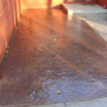 Stamped concrete walkway with a brown textured finish adjacent to a red exterior wall and a concrete path.