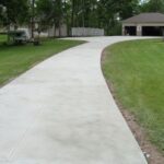 A long concrete driveway curves through a grassy yard toward a house with an open garage.